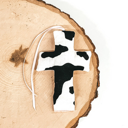 Cross shaped freshie that is a black and white cow print with light pink string. This is laying on a piece of wood that is on a white background.