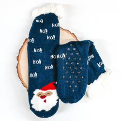 Navy socks that has Santa's face towards the bottom of the socks. These socks also have the word "HO!" on the ankle parts, has rubber grips on the bottom, and white fluff on the top of the socks. These socks are pictured laying on a piece of wood on a white background.