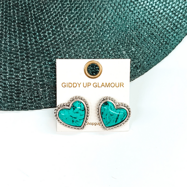 Turquoise, heart stone earrings with a silver backing and outline. These earrings are pictured on a white earrings holder. Then pictured on a white and black background.
