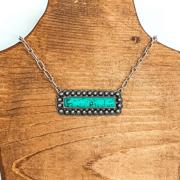 Silver chained necklace with silver rectangle pendant. The pendant has a center rectangle, turquoise stone that is outline with tiny, silver conchos. This necklace is pictured on a wood necklace holder on a white white background.