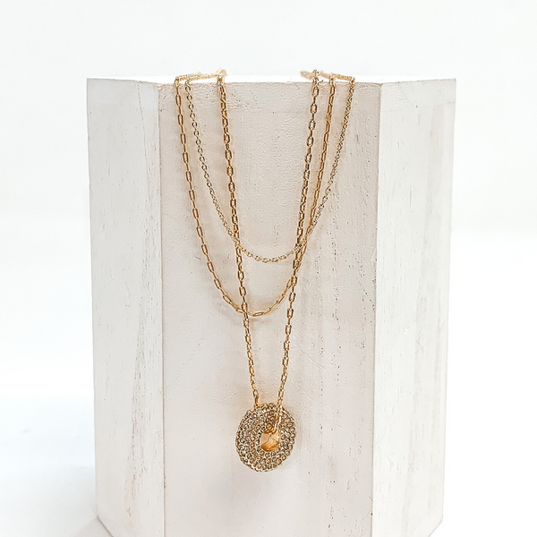 Gold, three stranded chained necklace with a round gold pendant. The pendant is covered in crystals. This necklace is pictured laying on a white block on a white background.