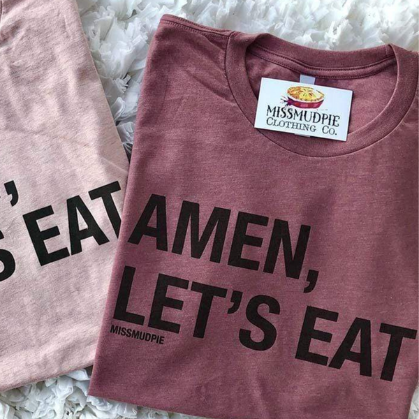 This mauve pink tee includes a crew neckline, short sleeves, and the words "AMEN, LET'S EAT" in an uppercase black block font. This tee is pictured here as a folded flatlay on white background. 