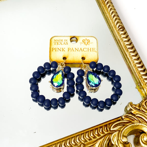 Circle drop navy beaded earrings with a hanging black ab teardrop crystal. These earrings are pictured on a wood earrings holder on a gold mirror on a white background. 