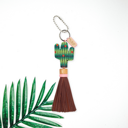 Centered in the middle of the picture is a beaded cactus charm with brown tassels. To the right of the charm is a palm leaf, all on a white background. 