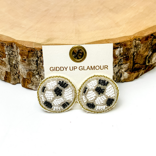 Pictured are beaded black and white soccer stud earrings. They are propped up on a piece of circle wood on a white background.