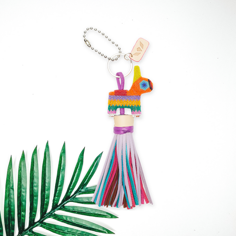 Centered in the middle of the picture is a beaded donkey charm with purple, pink, blue, and brown colored tassels. To the left of the charm is a palm leaf, all on a white background. 