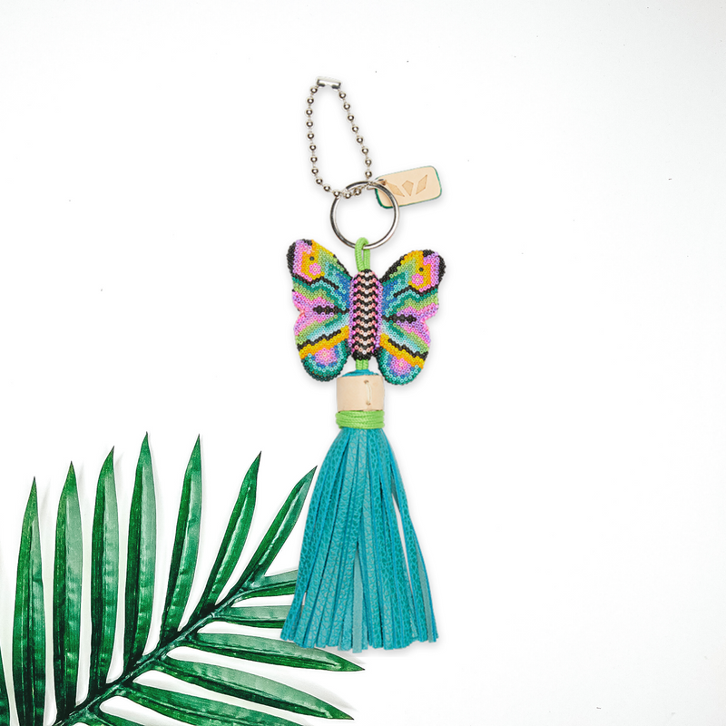 Centered in the middle of the picture is a butterfly charm with blue tassels. To the right of the charm is a palm leaf, all on a white background. 