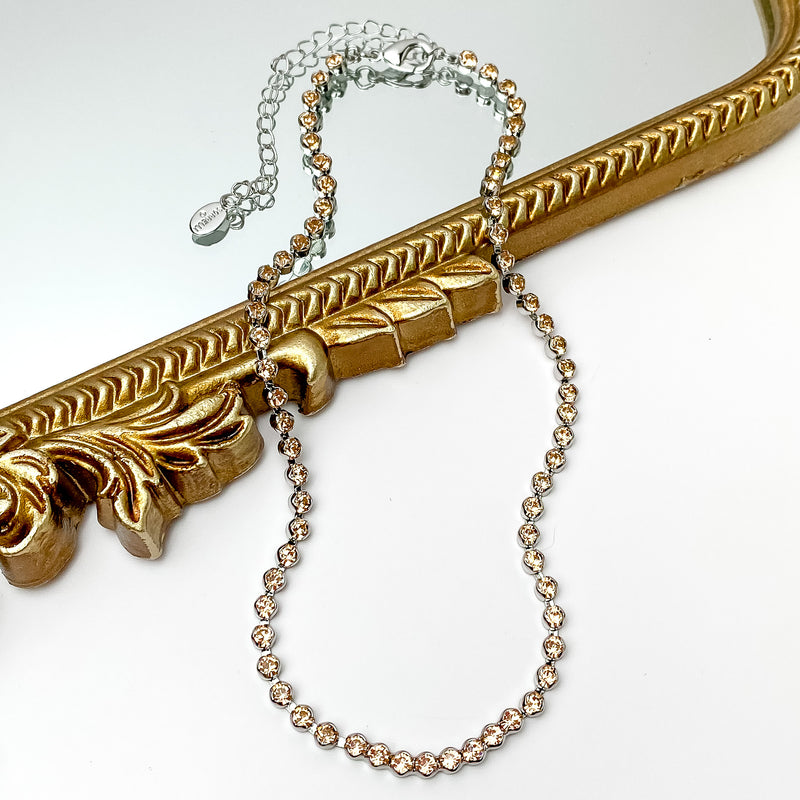 Pictured is a silver necklace with light beige crystals. This necklace is pictured partially on a gold mirror on a white background.