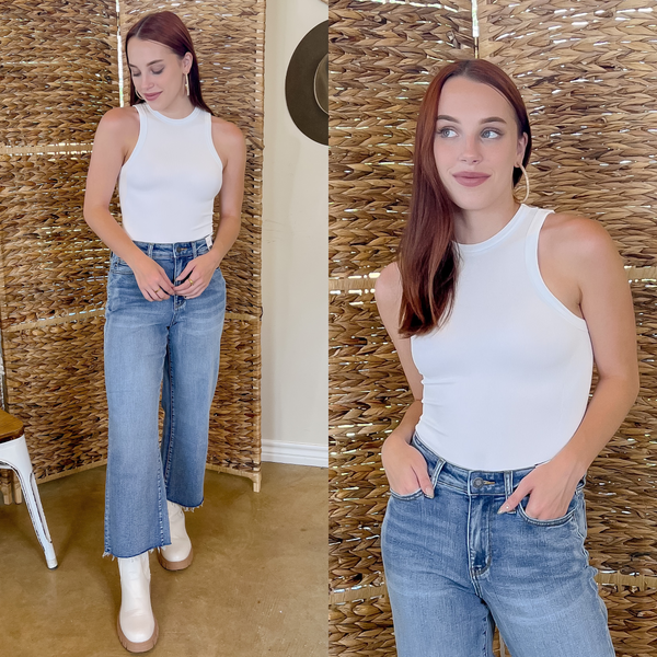 Model is wearing a white, tank top body suit and flare denim jeans. Model is also wearing white boots with rubber soles.