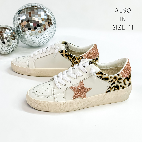 These tennis shoes are white in color with a leopard print back heel with rose gold glitter stars. These tennis shoes are pictured in front of disco balls on a white background.