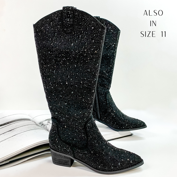 Pictured are black cowboy boots with black crystals covering the entire boot. These boots also include a black heel and sole. These boots are pictured on front on a white background with one boot resting on an open book.