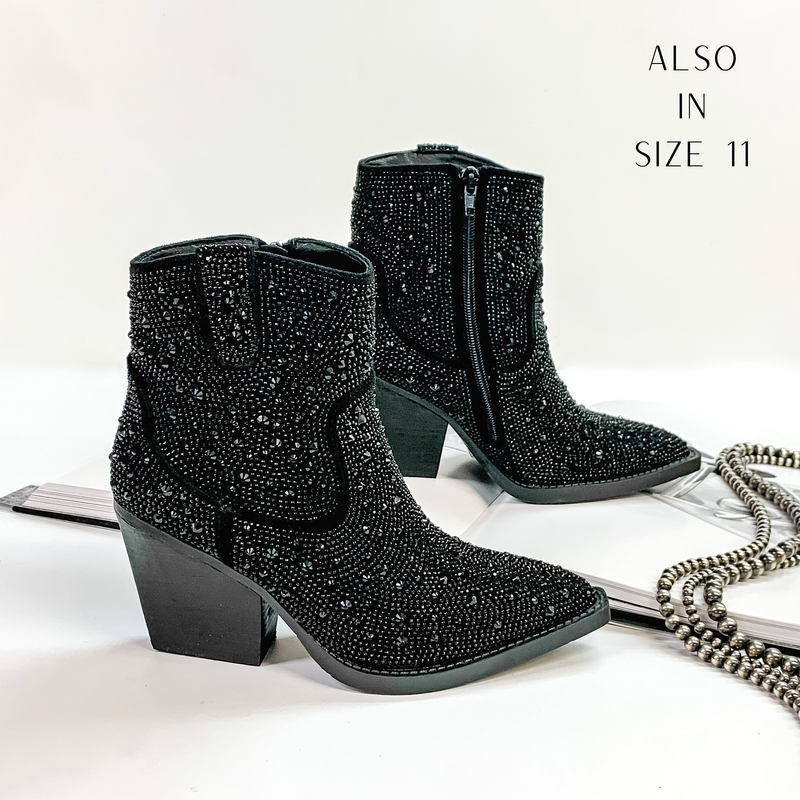 Pictured are black cowboy booties with black crystals covering the entire boot. These boots also include a black heel and sole. These boots are pictured on front on a white background with one boot resting on an open book.