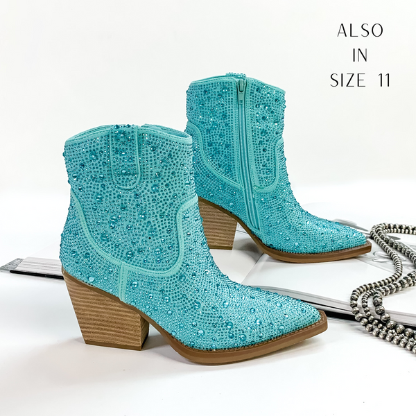 Pictured are black cowboy booties with turquoise crystals covering the entire boot. These boots also include a tan heel and sole. These boots are pictured on front on a white background with one boot resting on an open book.
