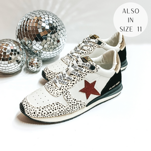 White tennis shoes with a cheetah print, gold, and black patches throughout the shoe. These shoes also have a rust red star emblem on the side of the shoe. These shoes are pictured on a white background with disco balls on the left hand side.