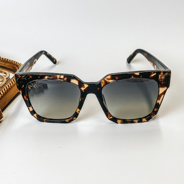 A pair of tortoise print frame sunglasses with gradient square lenses in black. These sunglasses are pictured on a white background with gold jewelry.