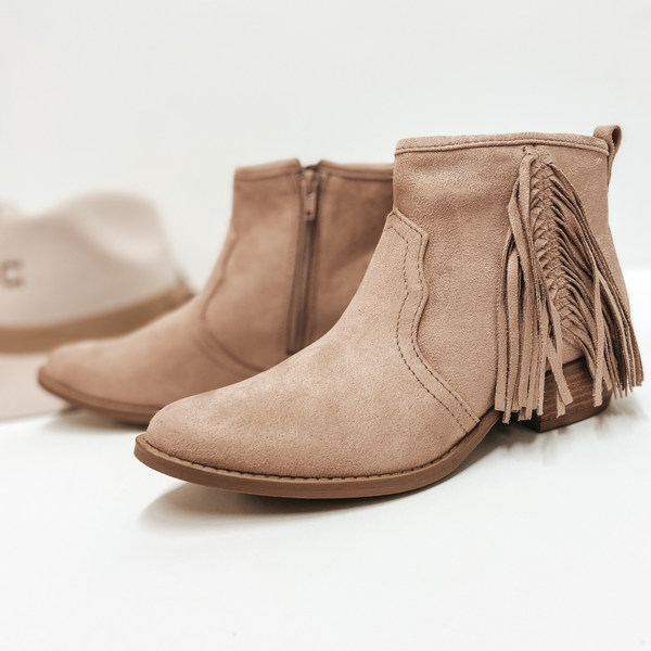 A pair of light brown suede booties with fringe on the side and a short heel. Pictured on white background with a felt rancher hat.