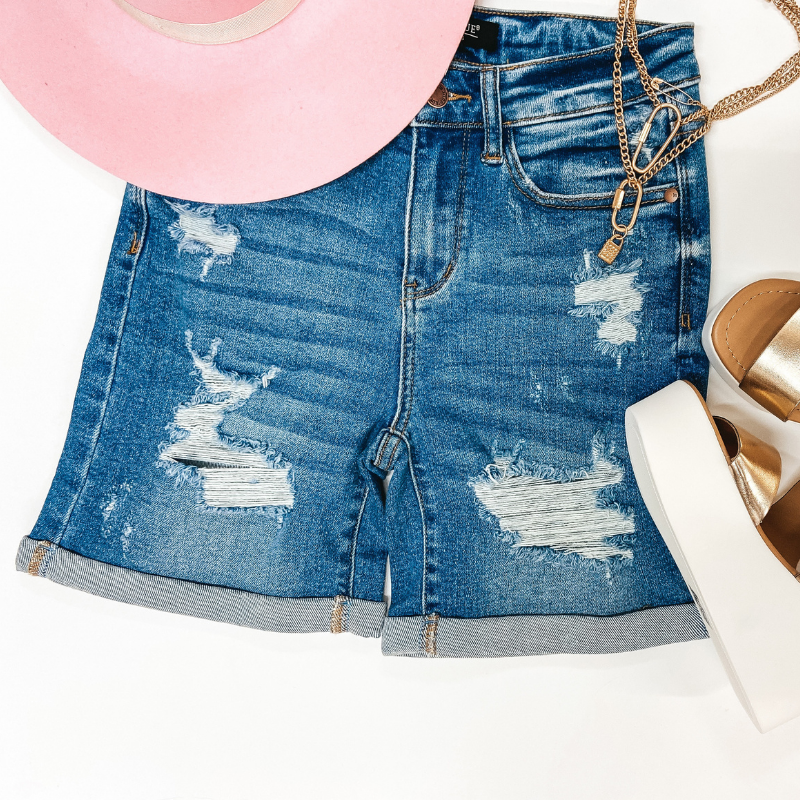 A pair of distressed mid thigh shorts with a cuffed hem. Pictured on white background with gold wedges, a gold necklace, and a pink hat.