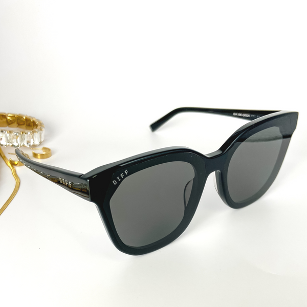 A pair of black cat-eye sunglasses with black lenses. These sunglasses are pictured on a white background with gold jewelry.