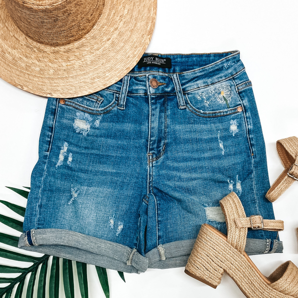 A pair of mid thigh shorts with dandelion embroidery and a cuffed hem. These shorts are pictured on a white background with a palm leaf and heeled sandals.