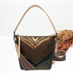 This bag has a smaller base with a wider top. This is a dark green bag that has an embroidered zebra print. The zebra print goes from an ivory color to a black color. This bag also has a striped handle and a wide tan leather strap. This bag is pictured on a white background with tan and brown pompous grass in the background. 