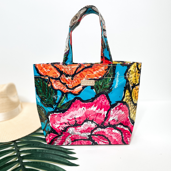 A small size bag that is a colorful floral print. Pictured on white background with a straw hat and palm leaf.