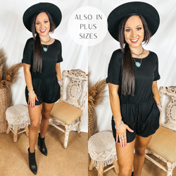 Model is wearing a black short sleeve romper with a drawstring waist. Model has it paired with a black hat, black booties, and silver jewelry.