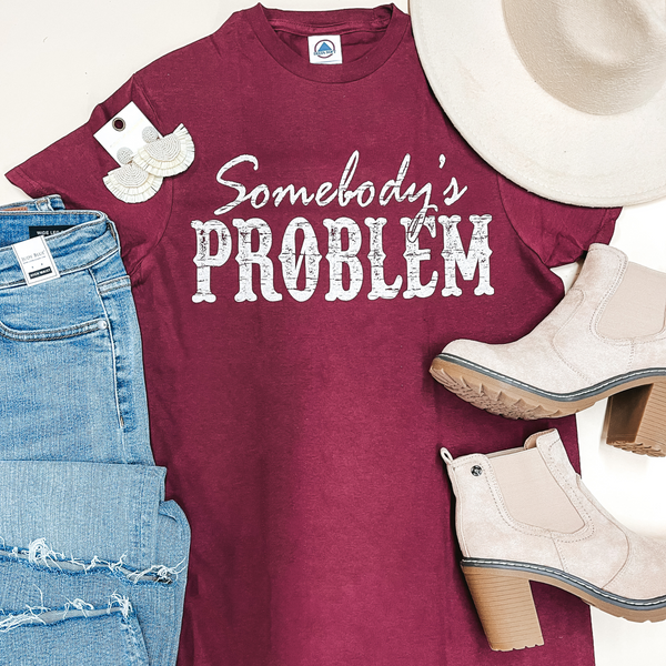 A maroon graphic tee that says "Somebody's Problem" in white writing. This tee is pictured on a white background with a beige hat, earrings, boots, and blue jeans.