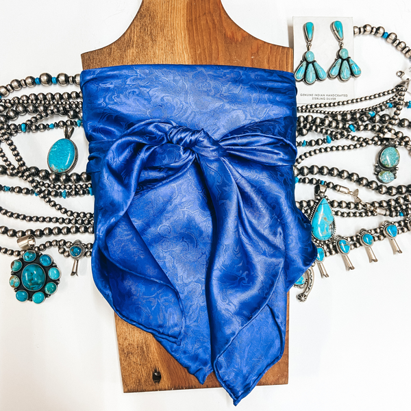 A jacquard print wild rag tied around a wooden display. Pictured on white background with sterling silver and turquoise jewelry.