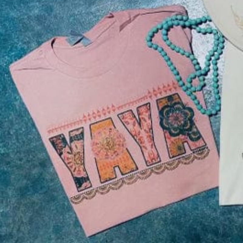 A light pink short sleeve tee with the word "YAYA" in the center. Each letter has its own unique pattern and color. There is a scalloped border below the text and a line border above. Item is pictured on a simple turquoise background.
