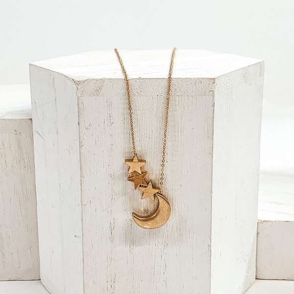 Simple gold necklace with gold moon charm and three gold star charms. This necklace is pictured on a white block on a white background.