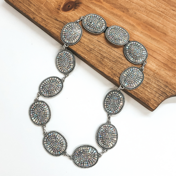 Silver, large oval concho hat band with covered in ab crystals. This hat band is pictured partially laying on a brown block on a white background.