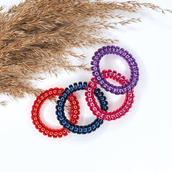 Pictured are four spiral scrunchies laying on top of each other in the colors purple, pink, blue, and red. These scrunchies are pictured on a white background with tan pompus grass in the top left corner.
