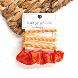A set of six hair ties, five spiral and one satin scrunchie in orange. There are three  solid color spiral hair ties and two transparent. These hair ties are on a white  cardboard piece with a thekitchenapproach logo, they are leaned up against a brown woven  plate and a white background.