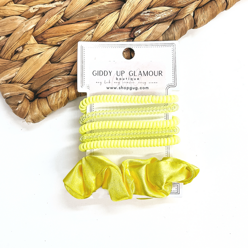 A set of six hair ties, five spiral and one satin scrunchie in yellow. There are three  solid color spiral hair ties and two transparent. These hair ties are on a white  cardboard piece with a thekitchenapproach logo, they are leaned up against a brown woven  plate and a white background.