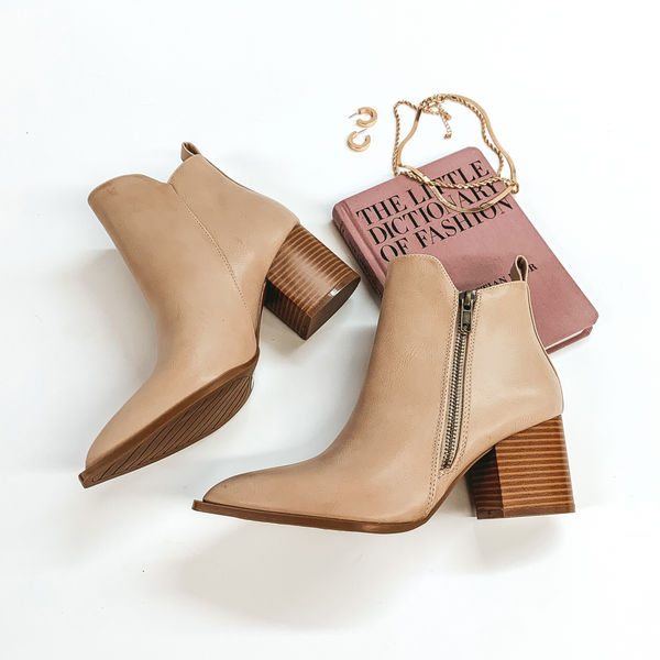 Ankle booties in beige with a side zipper. These booties also have a wooden heel. These booties are pictured on a white background with gold jewelry and a book.  