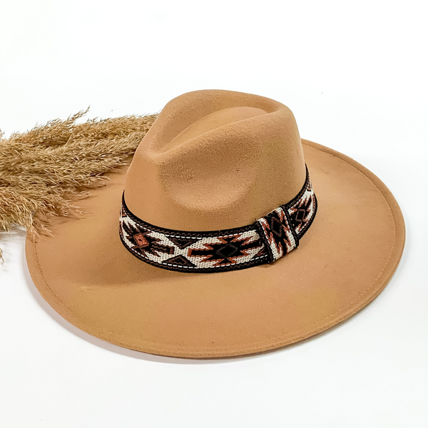 Taupe faux felt hat with a aztec print embroidered hat band. This hat is pictured on a white background with tan pompous in the background. 