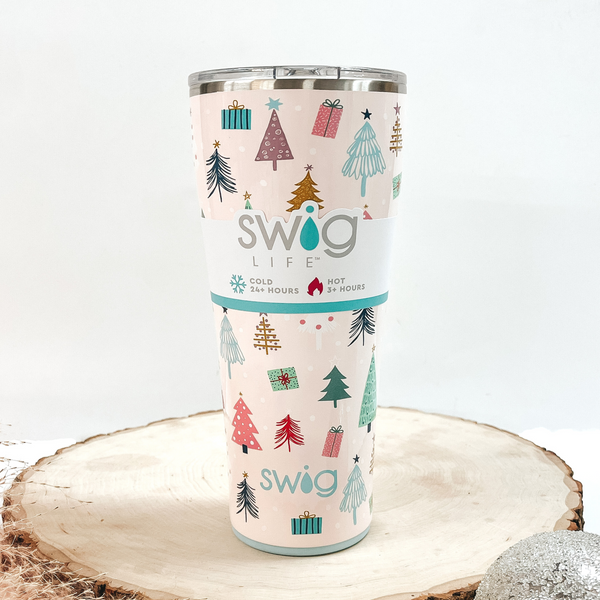Blush pink tumbler with multicolored tree and present print with a clear lid. This can cooler is pictured on a piece of wood on a white background.