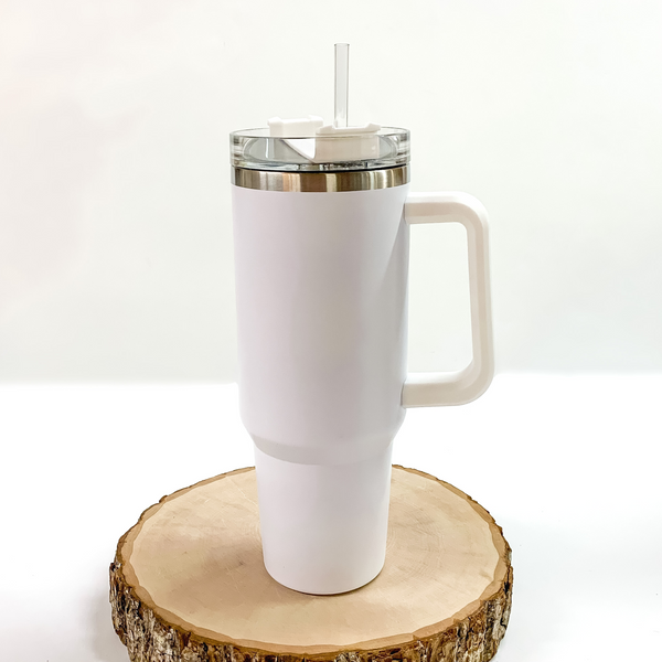 This is a white tumbler with a white handle. This tumbler also has a clear lid and straw. This tumbler is pictured standing on a piece of wood on a white background. 