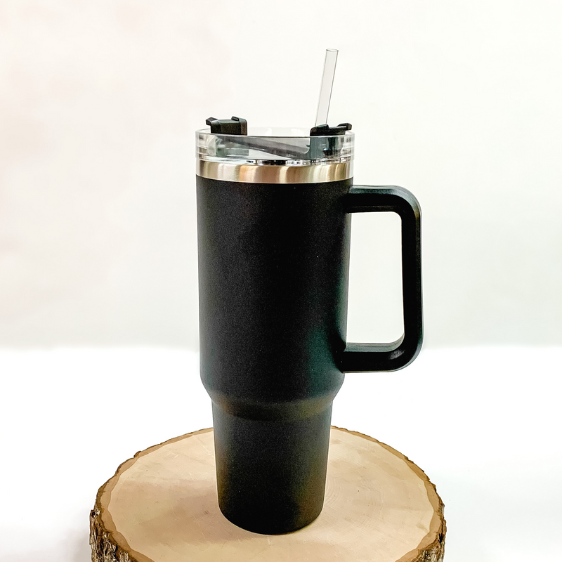 This is a black tumbler with a black handle. This tumbler also has a clear lid and straw. This tumbler is pictured standing on a piece of wood on a white background. 