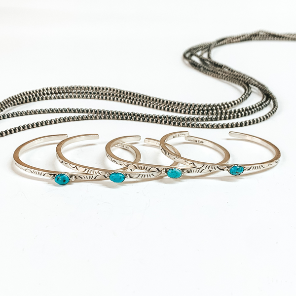 Four silver cuff bracelets that have detailed engraving and center turquoise stone. These bracelets are pictured laying on top of each other on a white background with silver beads behind them. 