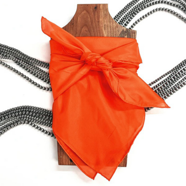 This is an orange silk wild rag, this wild rag is placed on a brown necklace board. This wild rag is taken on a white background and with silver Navajo pearls in the back as decor.