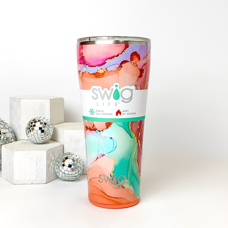 Pink and orange marble tumbler with a clear lid. This marble print includes shades of orange, pink, aqua, and gold glitter. This tumbler is pictured on a white background with white blocks and disco balls on the left side of the picture. 