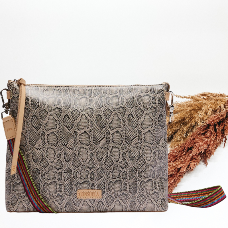 Pictured is a crossbody purse with a tan and black snake print all over design. This purse includes a thin, striped purse strap and a light tan tassel on the zipper. This purse is pictured on a white background with pompous grass on the right side of the picture.