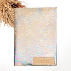 Pictured is a notebook cover with an iridescent multicolored design and a light tan Consuela patch on the bottom right corner. This notebook is pictured on a white background with pompous grass in the top left corner of the picture.