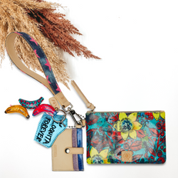 Blue pouch with red, yellow, and navy design with a navy wristelt that has a pink star print. This pouch also includes a card holder charm and some banana charms. This pictured on a white background with tan and brown pompous grass in the background.