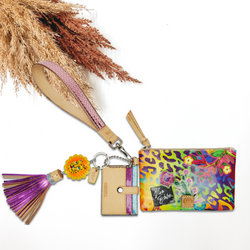 Multicolored leopard with different sticker pathces and a pink wristelt that has a woven design. This pouch also includes a card holder charm and a a lion head charm with purple tassels hanging from the bottom. This is pictured on a white background with tan and brown pompous grass in the background.