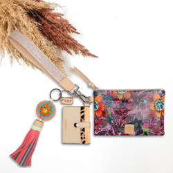Pink pouch with red, orange, and navy design with a silver glitter wristlet. This pouch also includes a card holder charm and a lion head charm that has a pink and blue tassel. This pictured on a white background with tan and brown pompous grass in the background.