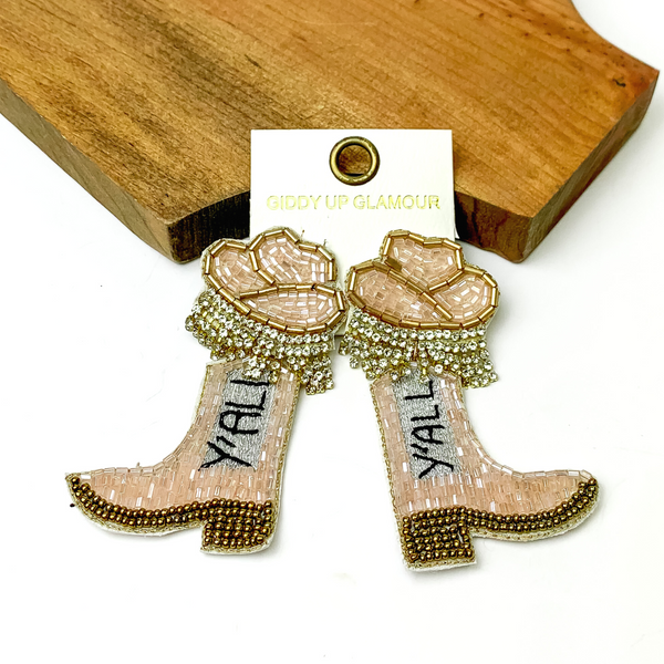 These earrings are pale pink beaded hat post back earrings with a pale pink beaded boot pendant. These earrings include gold beaded outline, crystal fringe on the bottom of the hat, and the word "Y'ALL" stitched in black. These earrings are pictured on a white background with a brown block at the top of the picture.