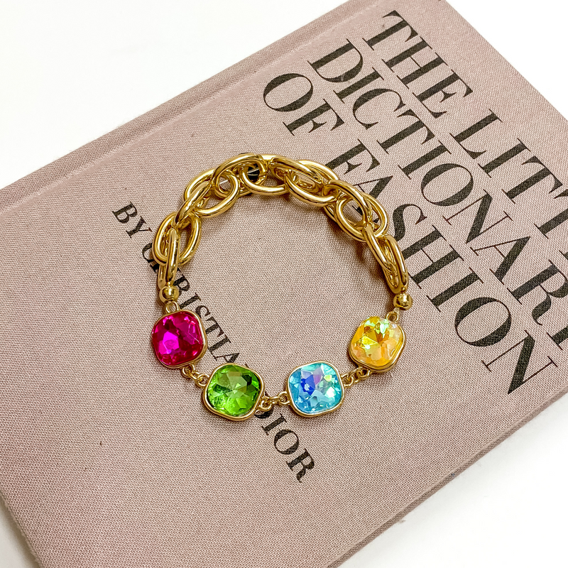 Gold Chain bracelet with four cushion cut crystals. These crystals come in fuchsia pink, yellow, blue, and green. This bracelet is pictured on a mauve colored book on a white background. 