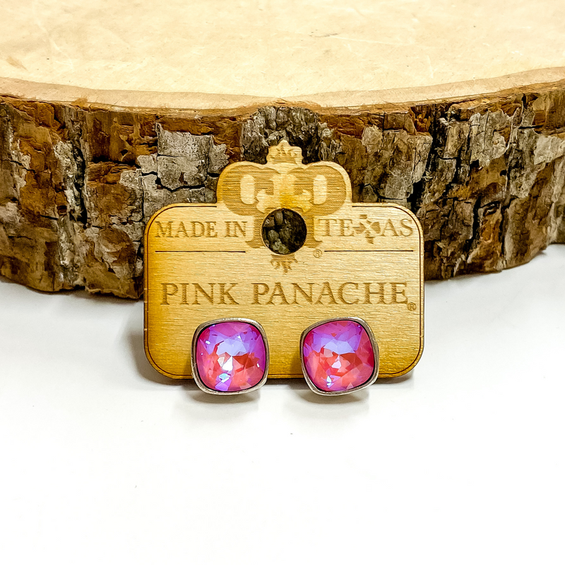 A pair of silver, square stud earrings with pink lotus delight cushion cut crystals. These earring are pictured on a wood holder on a white background with a piece of wood in the background.